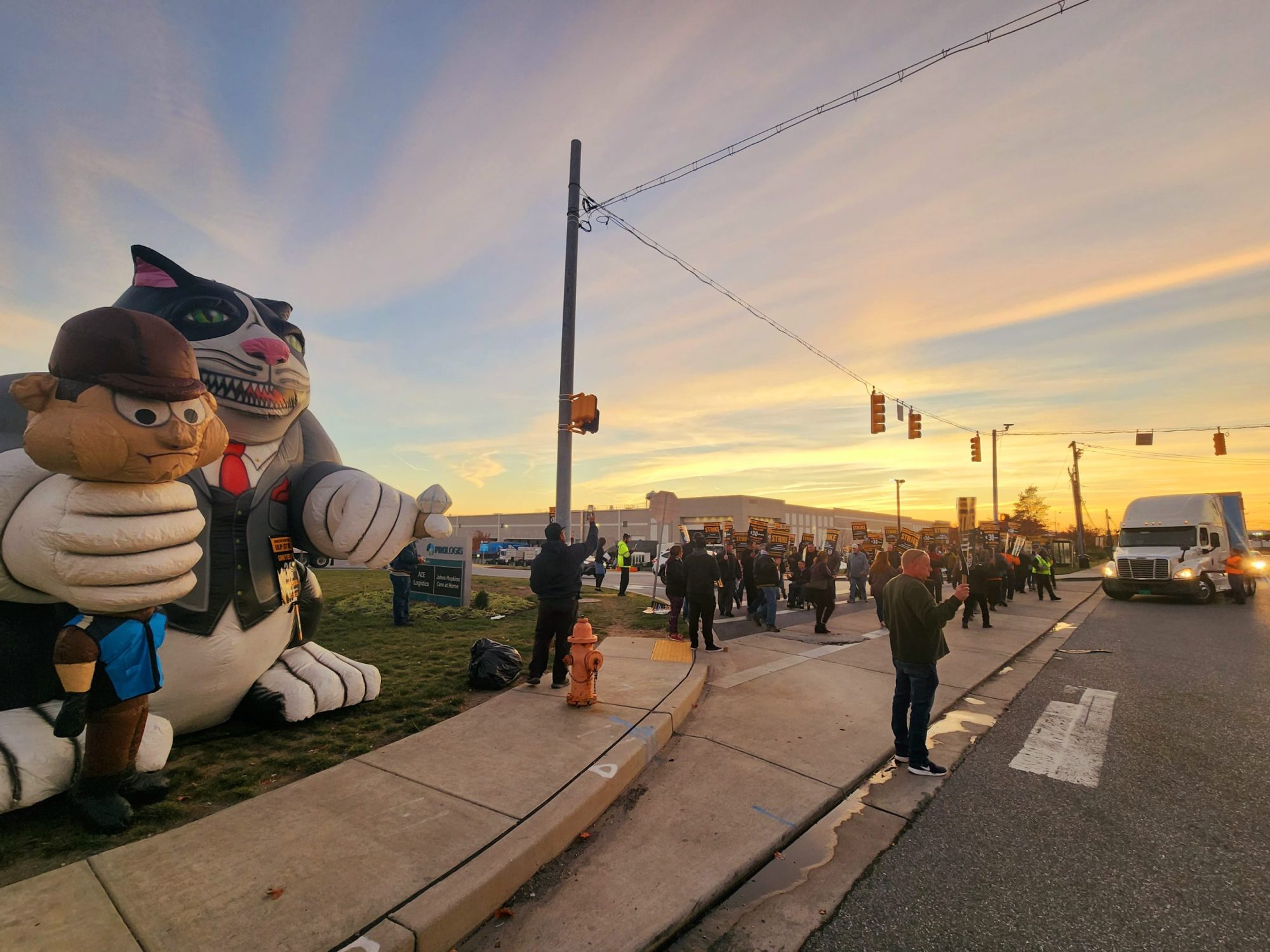 A group of dozens of Teamsters and local community members walk a picket line at sunset outside Amazon's BWI5 warehouse in Baltimore on Wednesday, Nov. 8, 2023. On the corner of the intersection stands a "Fat Cat" blowup doll. Photo by Maximillian Alvarez.