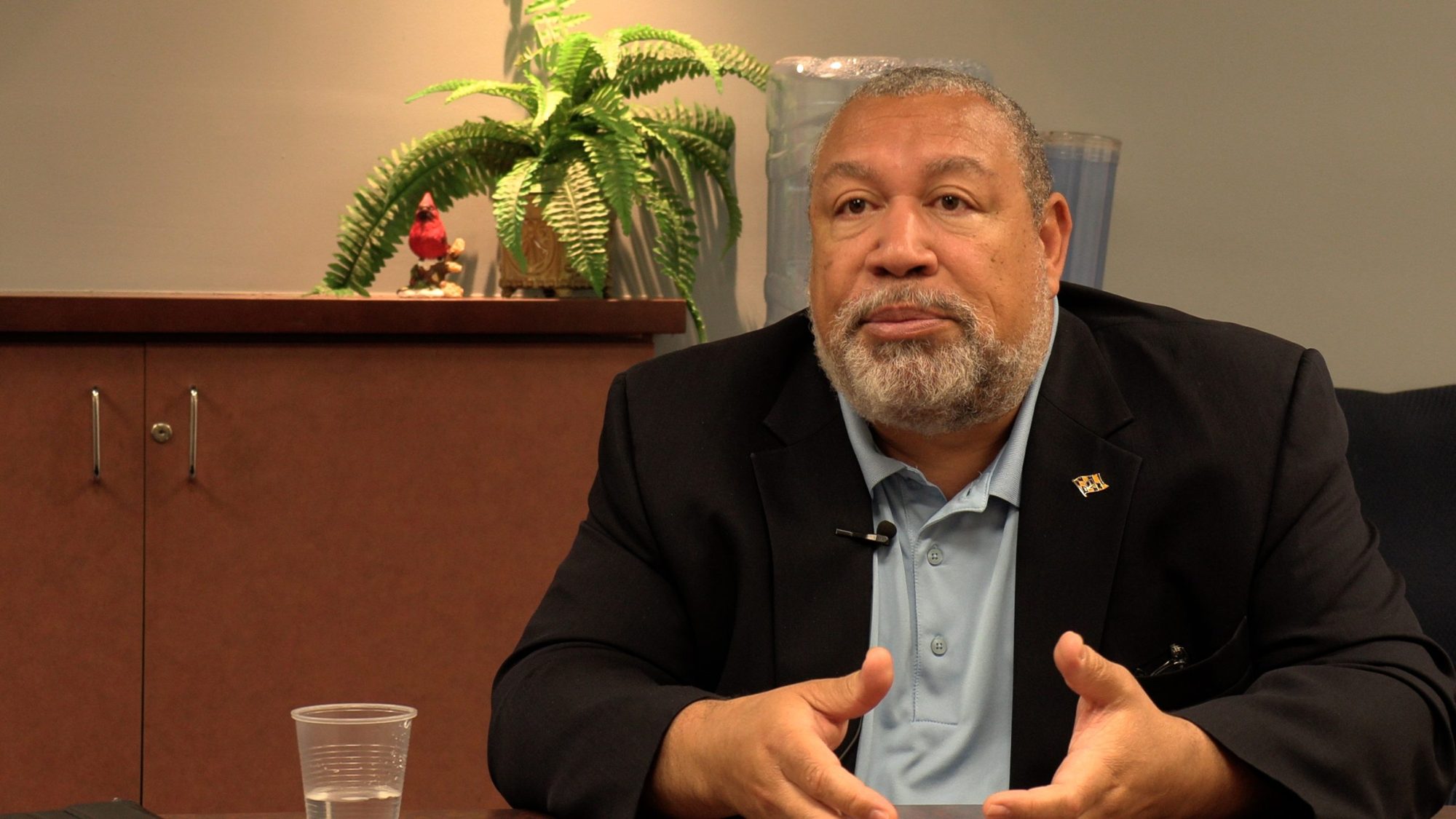 Bill Henry sits at a desk and explains something during an interview. He is an older Black man with a light beard, wearing a dark suit and button up shirt with no tie.