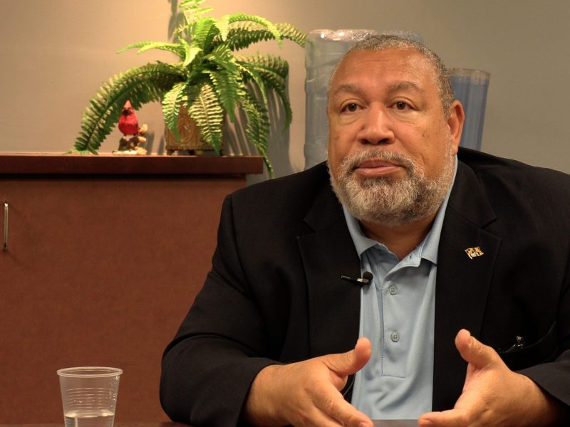 Bill Henry sits at a desk and explains something during an interview. He is an older Black man with a light beard, wearing a dark suit and button up shirt with no tie.