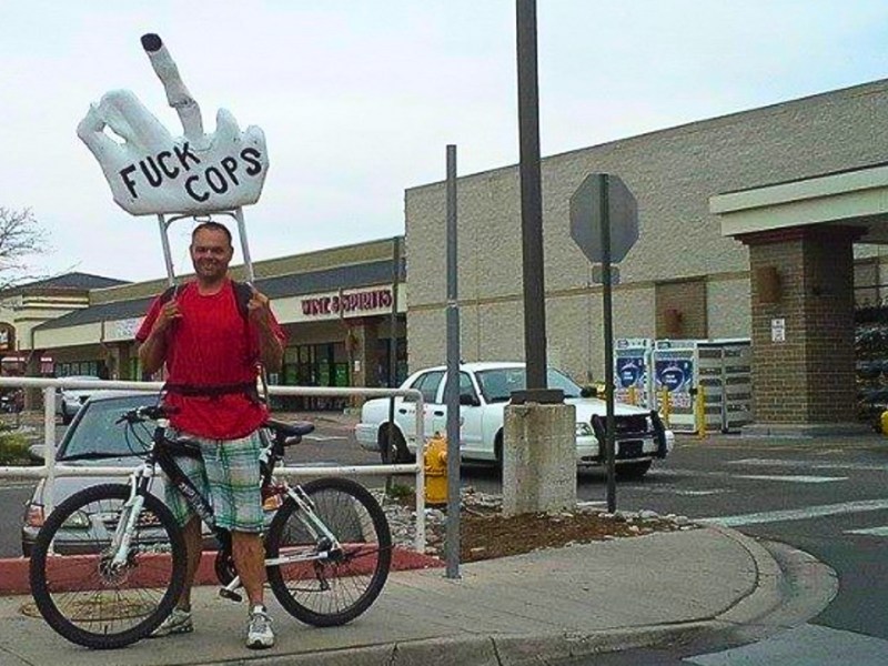 Photo of Eric Brandt exercising his First Amendment rights in Denver, Colorado, posing on a bicycle holding a giant hand over his head, which is pointing a middle finger and has "FUCK COPS" written on it. Photo courtesy of Eric Brandt.