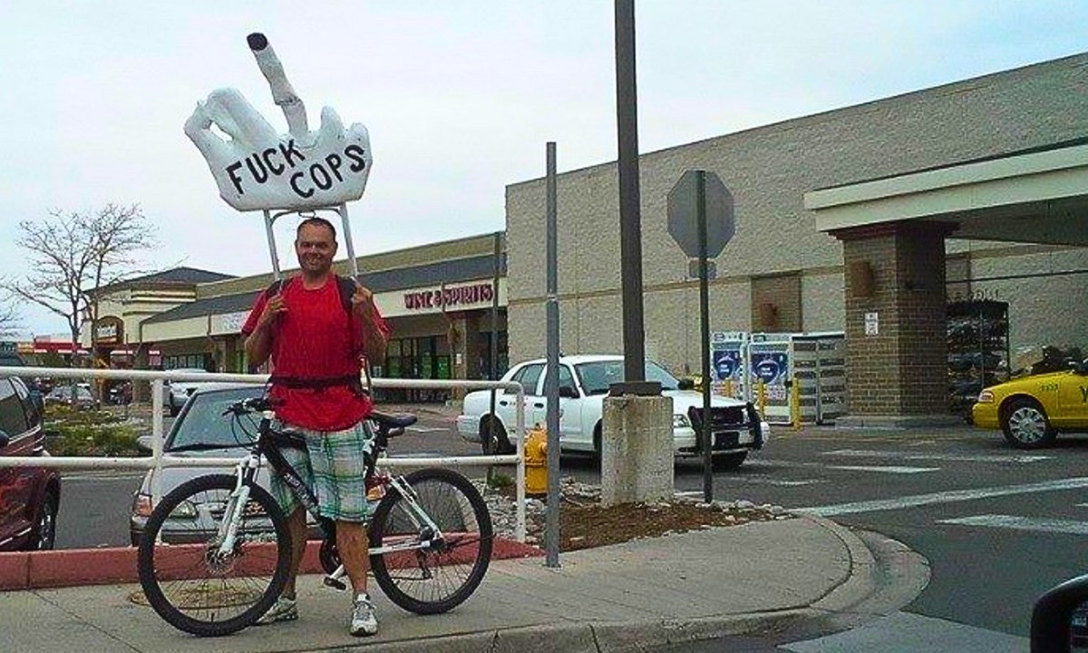 Photo of Eric Brandt exercising his First Amendment rights in Denver, Colorado, posing on a bicycle holding a giant hand over his head, which is pointing a middle finger and has "FUCK COPS" written on it. Photo courtesy of Eric Brandt.