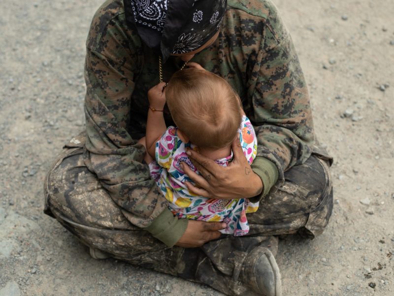 A person in camo gear sits criss-crossed on the ground, cradling a baby.