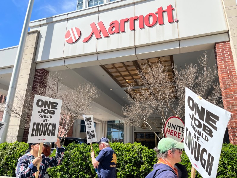 Low angle shot that showing three picketers marching in beneath the Marriott logo. The picketers carry signs that say "ONE job should be enough."