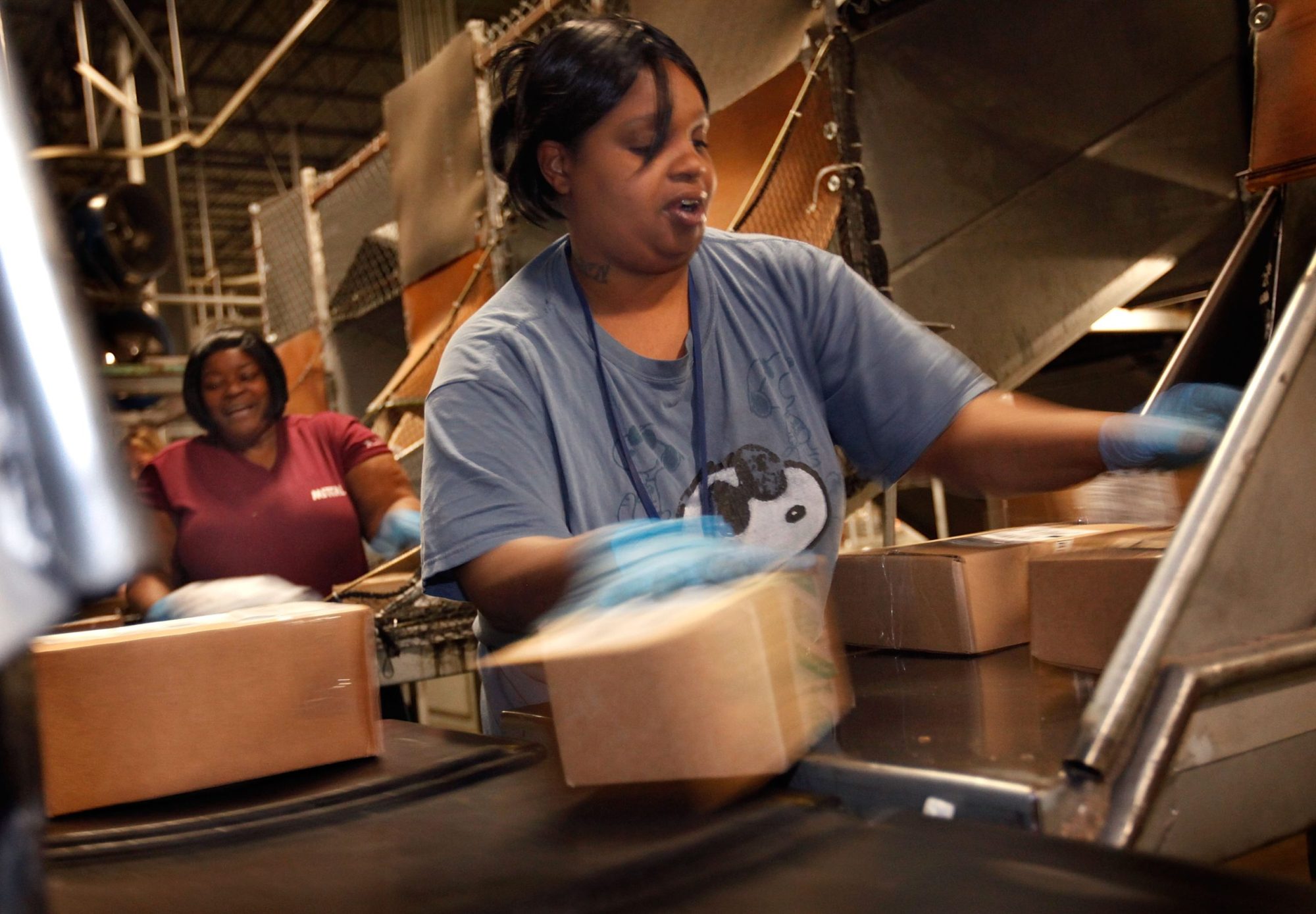 Two Black women stand at a conveyer belt where packages of various sizes are coming down a series of chutes and tubes. The woman in the foreground is wearing a blue shirt, and her arms are somewhat blurred from the speed of her sorting the packages. Her mouth is open mid-speech. In the background, her coworker laughs, apparently responding to something she has heard.