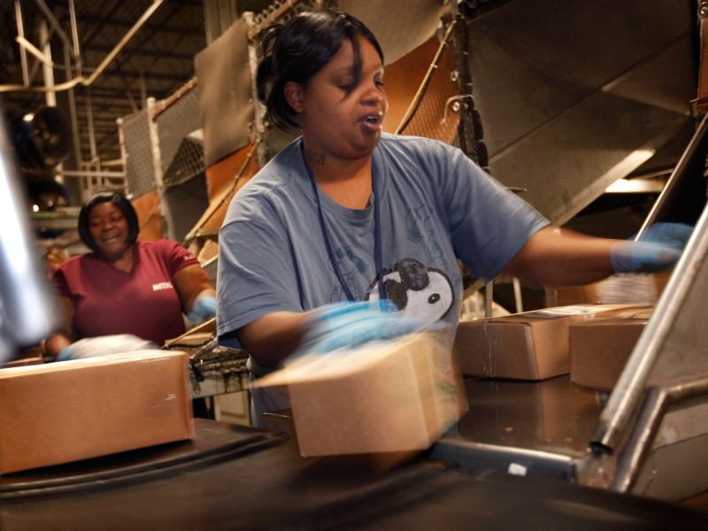 Two Black women stand at a conveyer belt where packages of various sizes are coming down a series of chutes and tubes. The woman in the foreground is wearing a blue shirt, and her arms are somewhat blurred from the speed of her sorting the packages. Her mouth is open mid-speech. In the background, her coworker laughs, apparently responding to something she has heard.