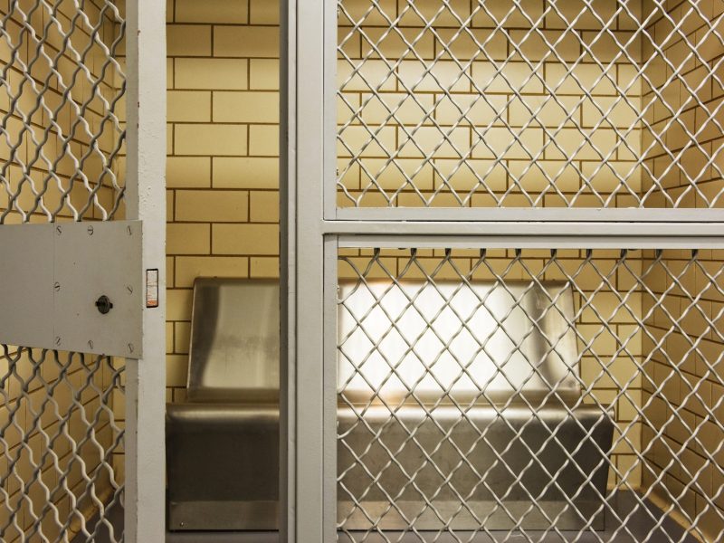 A small tiled cell with a mesh wire door and a metal bunk.