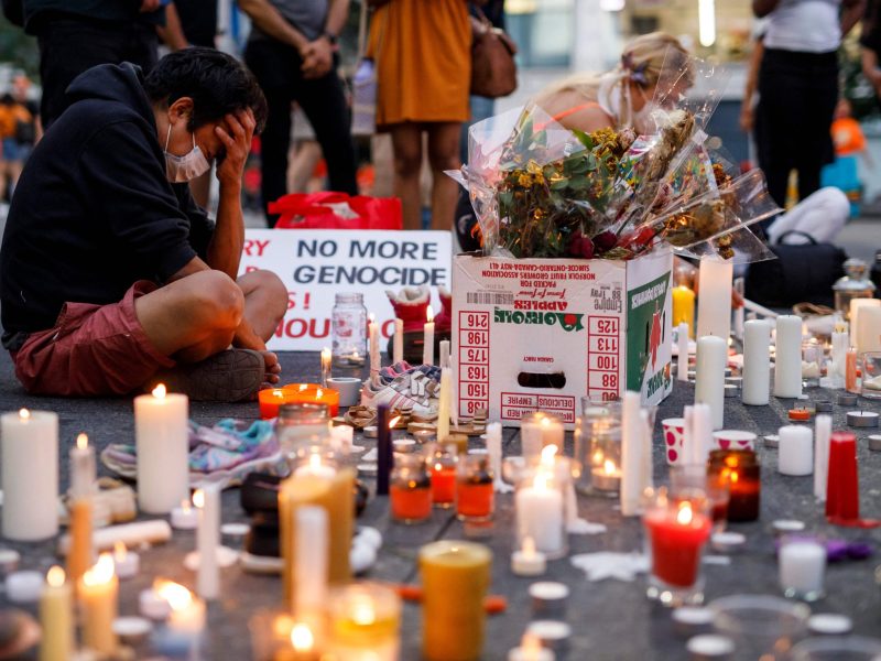 A man sits on the pavement holding his head in his hands. He is surrounded by lit candles in an impromptu vigil.