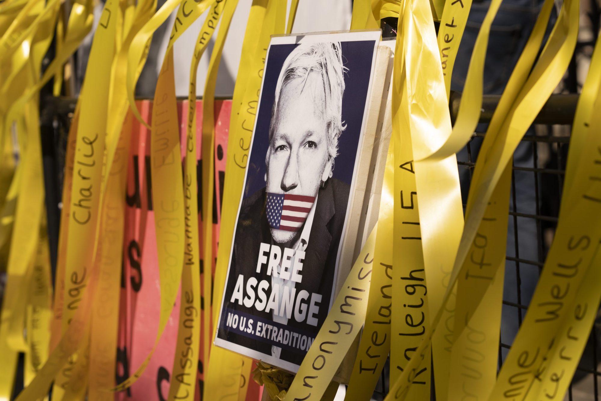 A poster of Julian Assange with his mouth covered by the US flag. Around the poster are yellow streamers demanding his release.