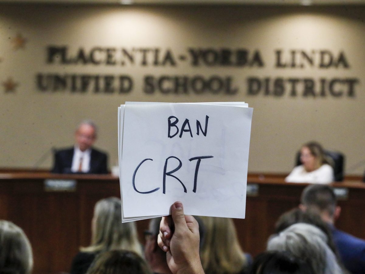 Hand holding a sign up in front of a school board that reads 'Ban CRT'