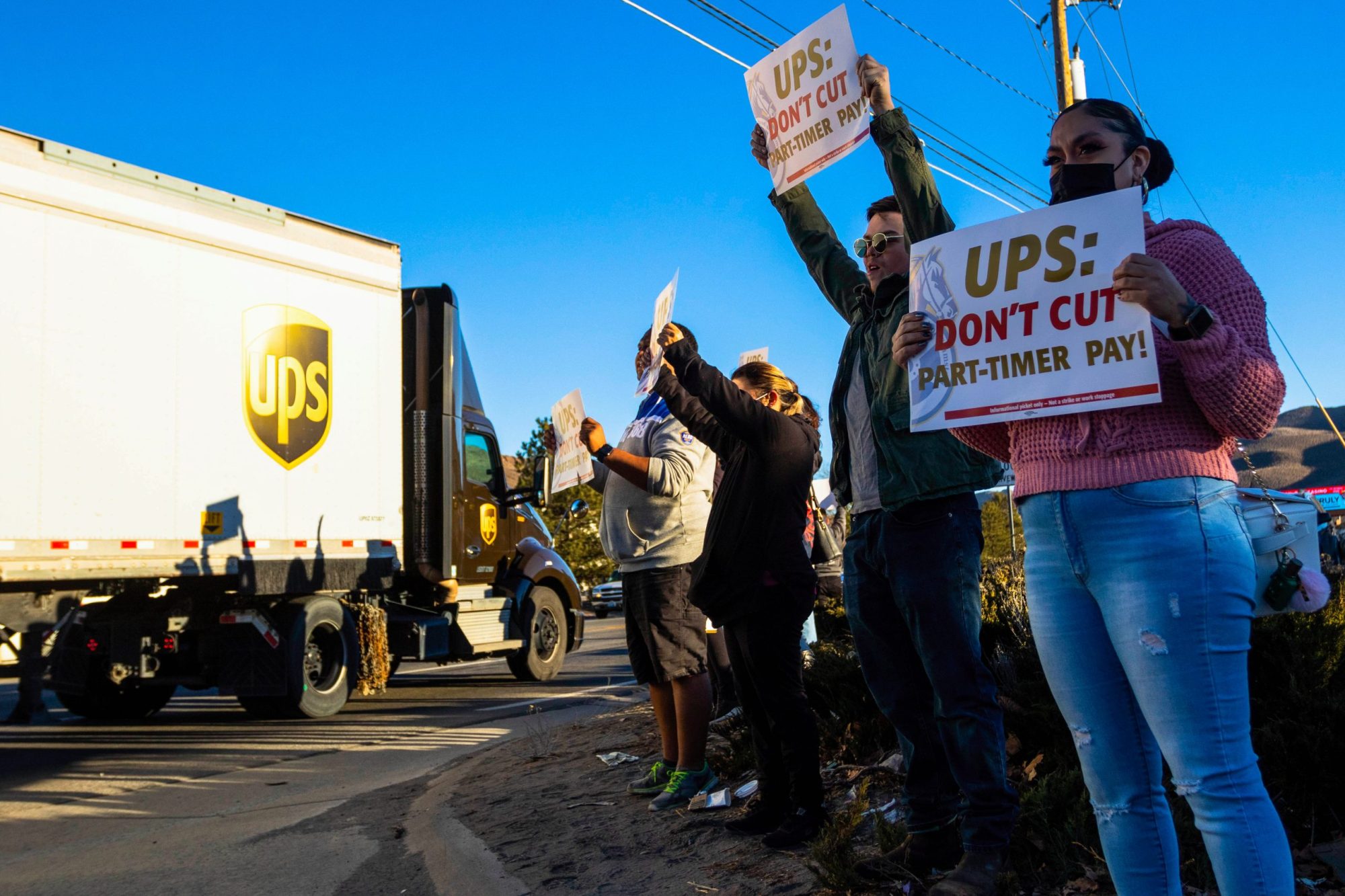 Four protesters holding placards stand at the curved dirt shoulder of a road as a massive 18-wheeler UPS semi-truck with the logo displayed on the side drives past. The protesters' signs say "UPS: Don't cut part-timer pay!"