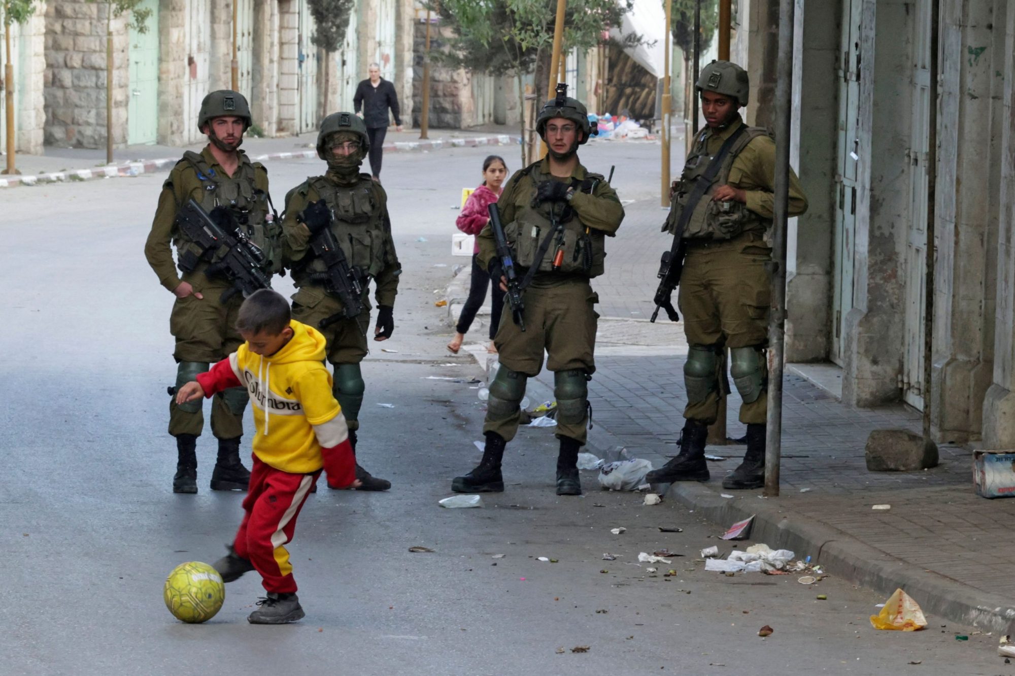 A small boy dribbles a soccer ball on a street as four armored Israeli soldiers carrying automatic weapons stand behind him and watch.