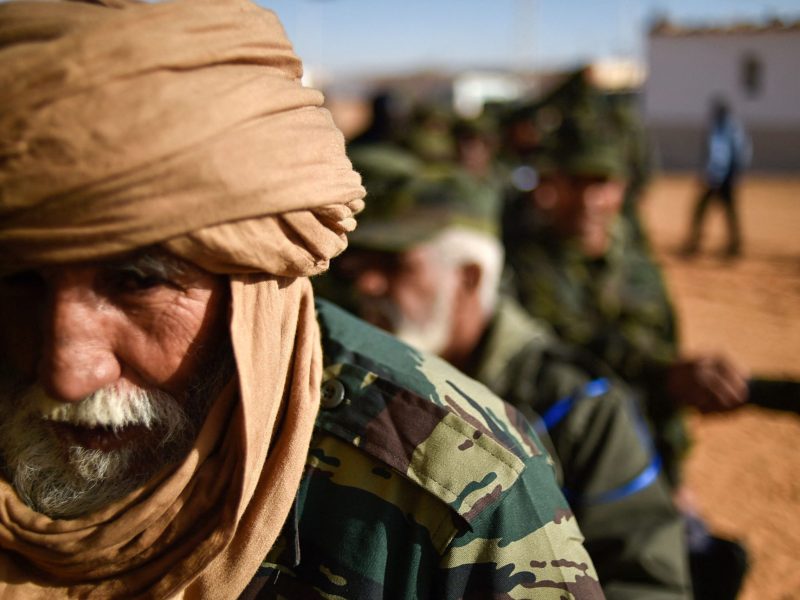 Close up of the face of an older Sahrawi man. He is bearded and his head is covered by cloth. He is wearing military fatigues, and appears to be seated in a desert environment. Behind him, other men dressed similarly can be seen sitting in a row.