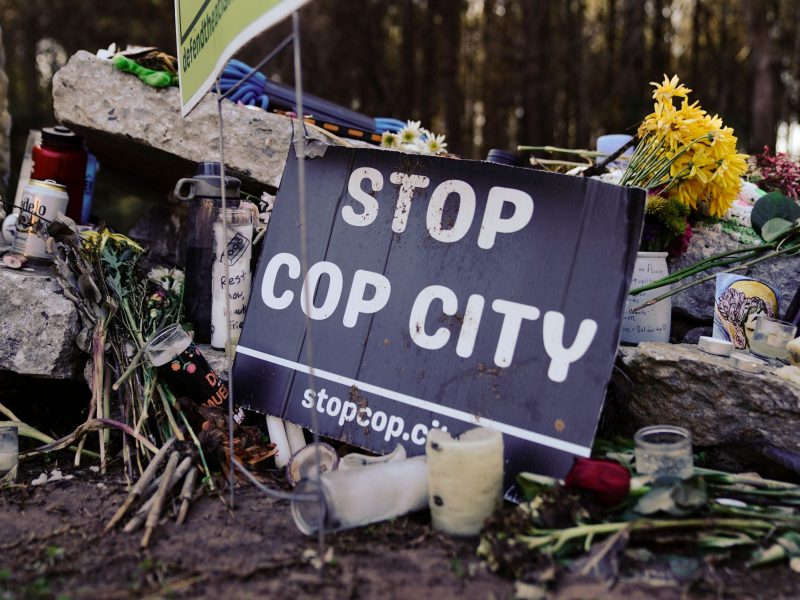 A "Stop Cop City" sign is positioned in front of a makeshift memorial featuring flowers and offerings.