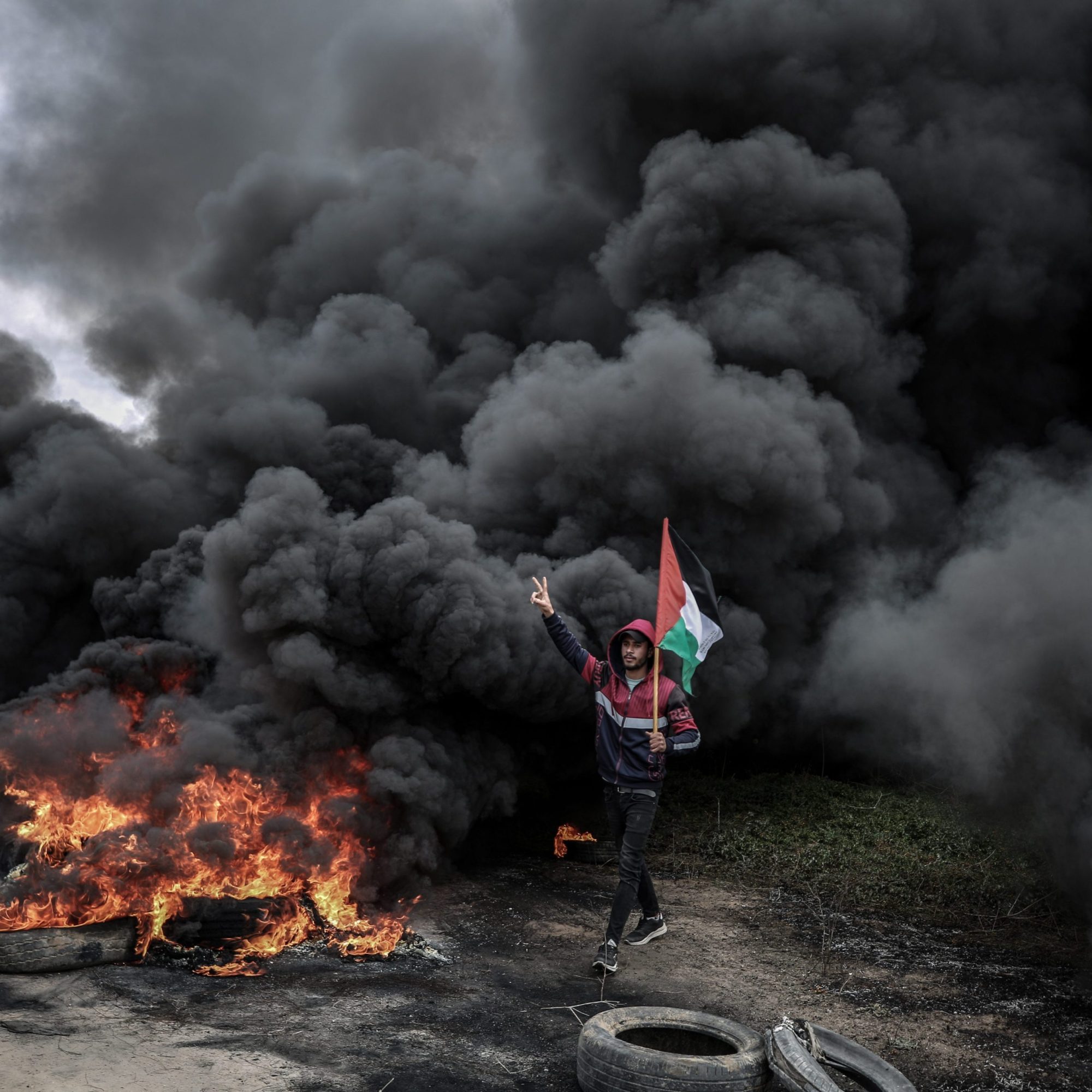 A young Palestinian man waves his flag in front of a thick column of black smoke