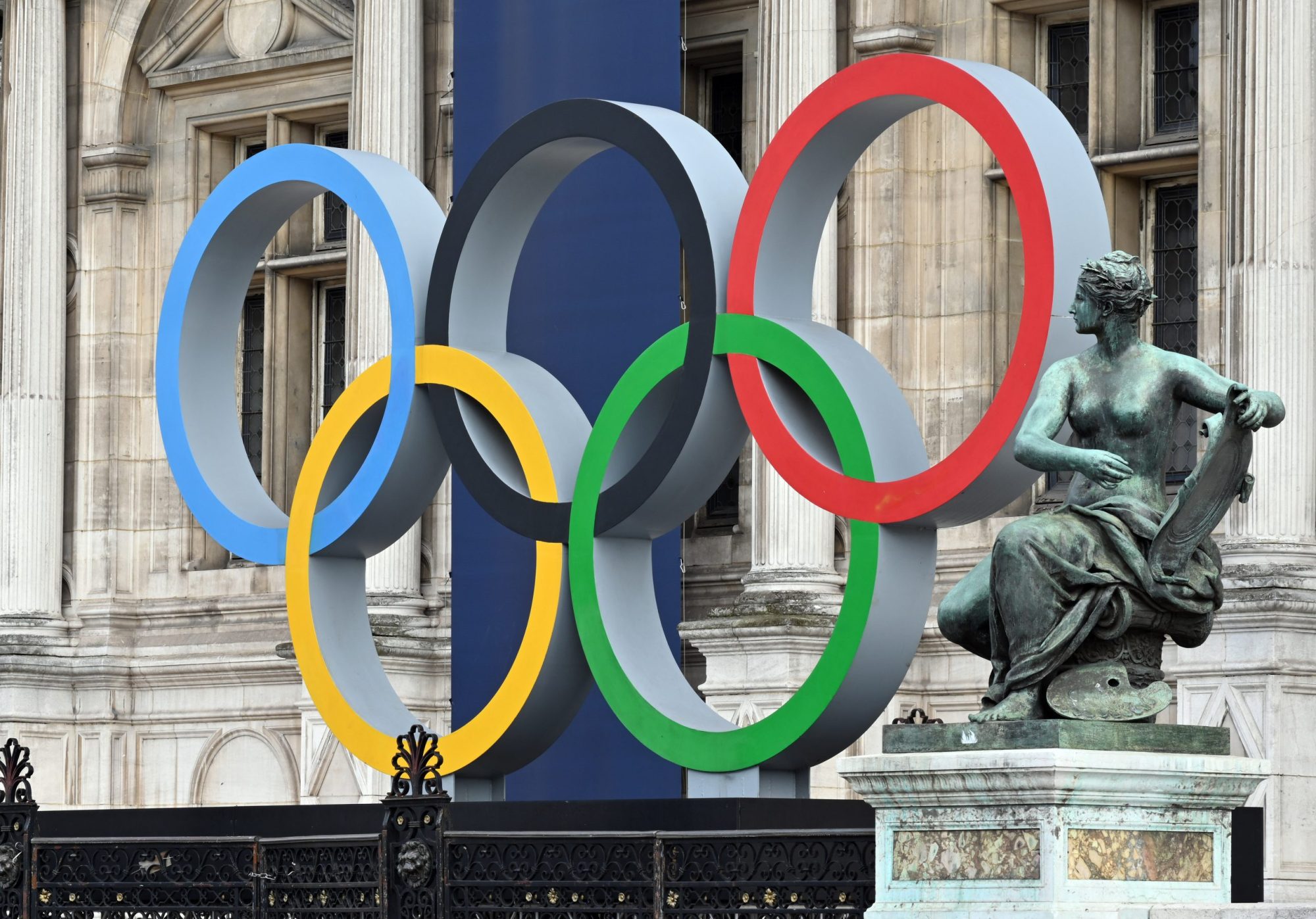 The five olympic rings displayed in front of a copper statue of an angel