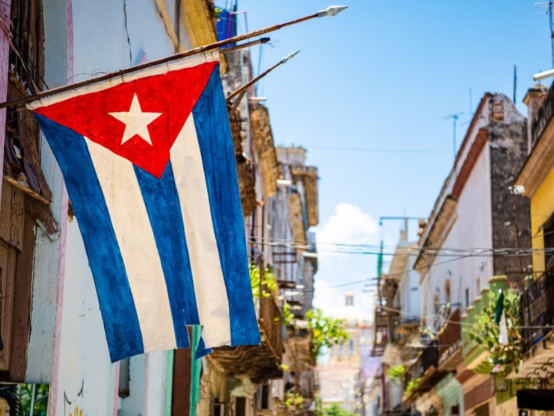 Closeup image of a small Cuban flag hanging at the entrance of a narrow street flanked by colorful buildings