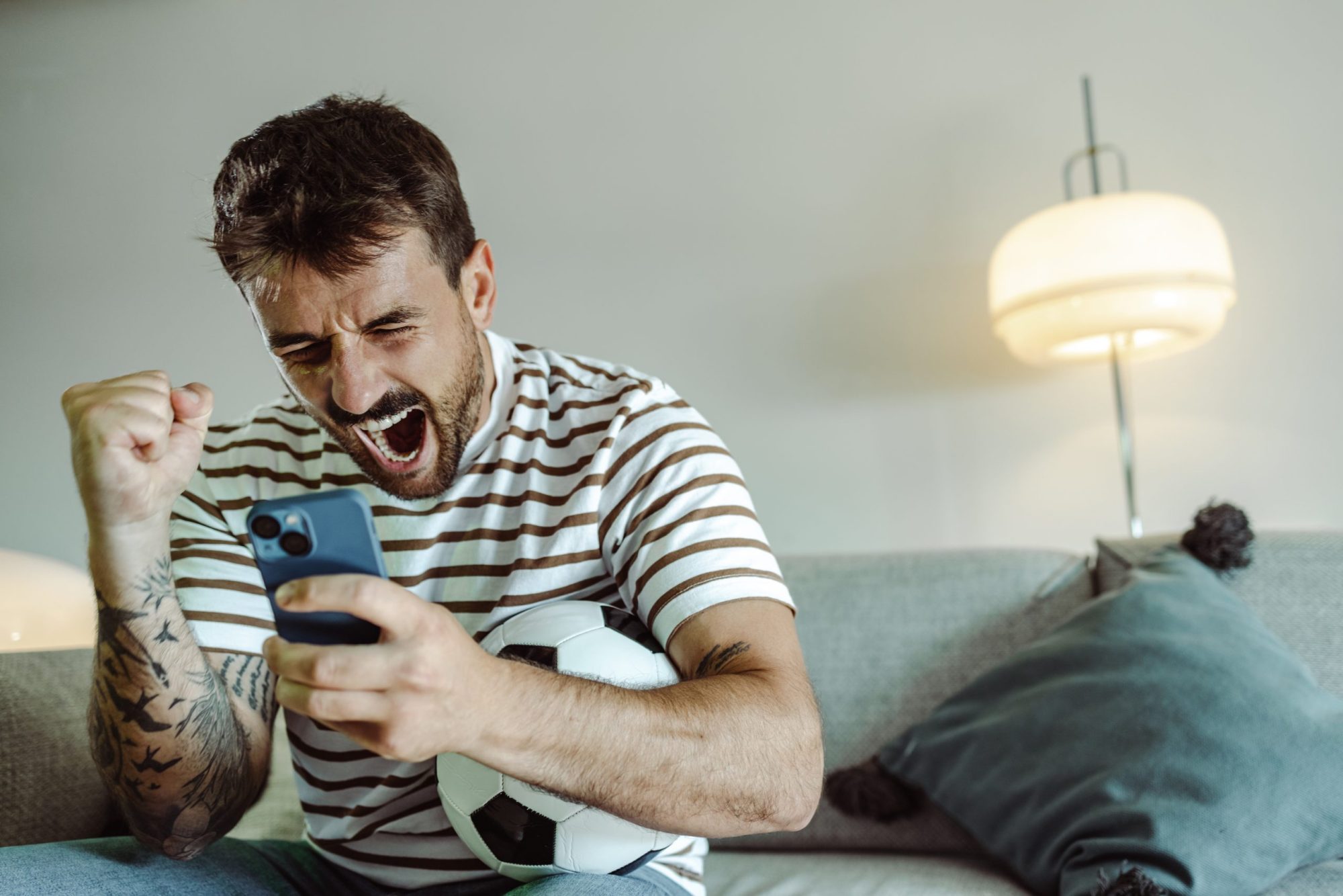 Excited young man watching sports game and cheering for his team, He is holding mobile phone and he is happy about winning the bet.