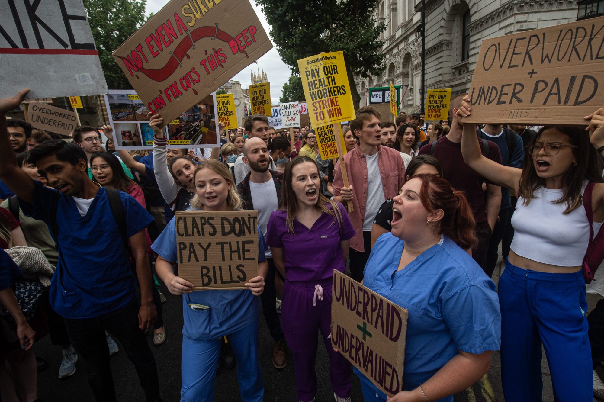 Ground-level shot of a crowd of protestors, many of them wearing medical scrubs. The signs they bear display slogans like, "Claps don't pay the bills," and "Underpaid + undervalued."