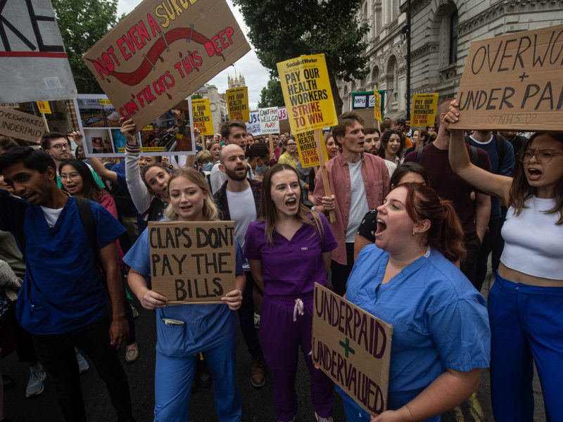 Ground-level shot of a crowd of protestors, many of them wearing medical scrubs. The signs they bear display slogans like, "Claps don't pay the bills," and "Underpaid + undervalued."