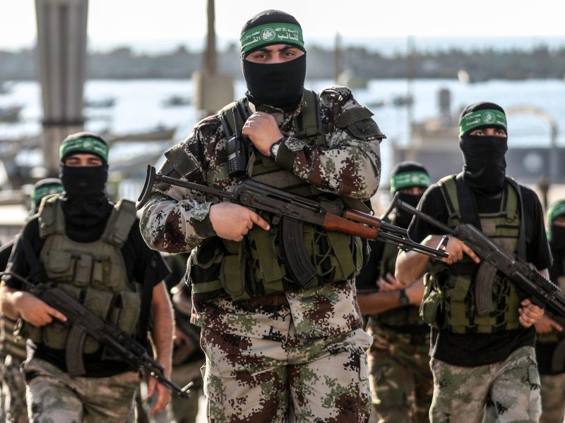 Three masked, uniformed fighters of the Al-Qassam brigades carrying automatic weapons. They are wearing camouflage fatigues, black ski masks, and the iconic green headband of Hamas fighters.