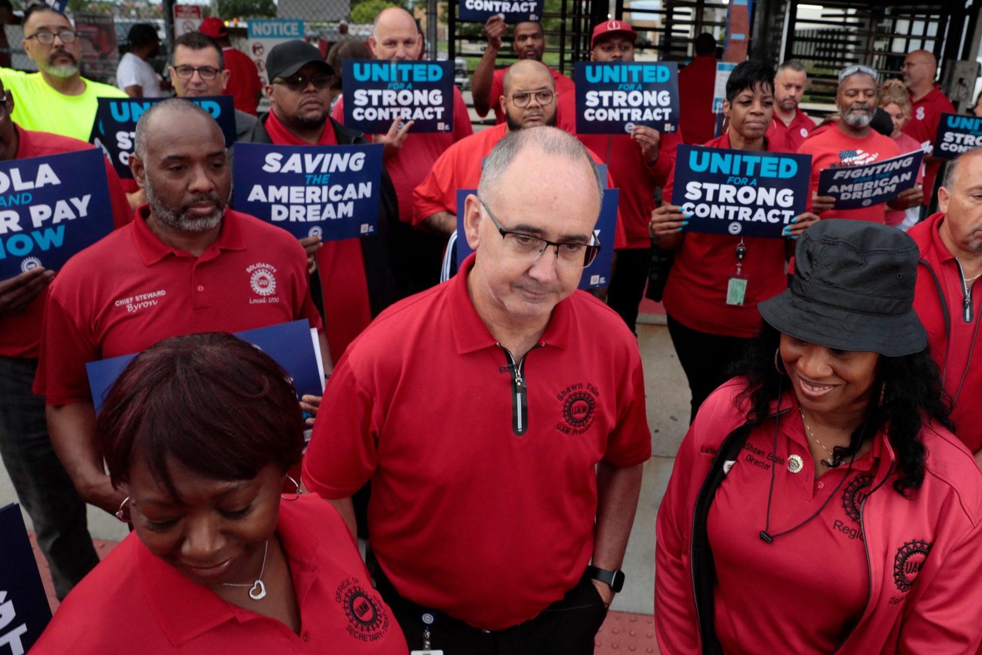 A group of UAW workers in matching red shirts stand together in a group, holding signs demanding a fair contract.