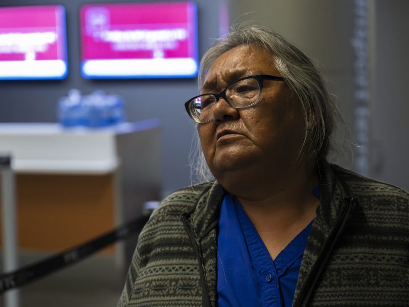 An older Indigenous woman sits inside an office. She is wearing glasses, has long grey hair, and a bright blue shirt. She has a grieving expression on her face.