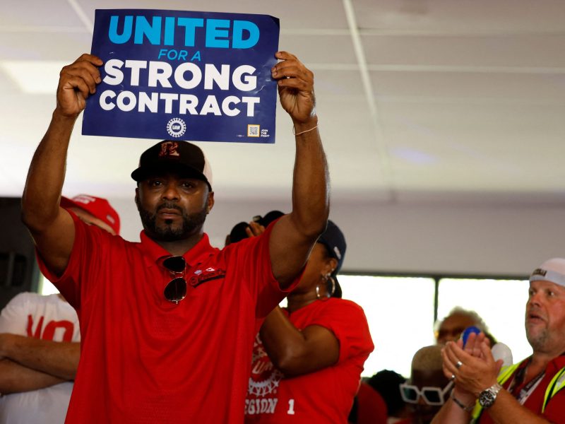 A worker in a red shirt holds up a sign that reads "United for a Strong Contract"