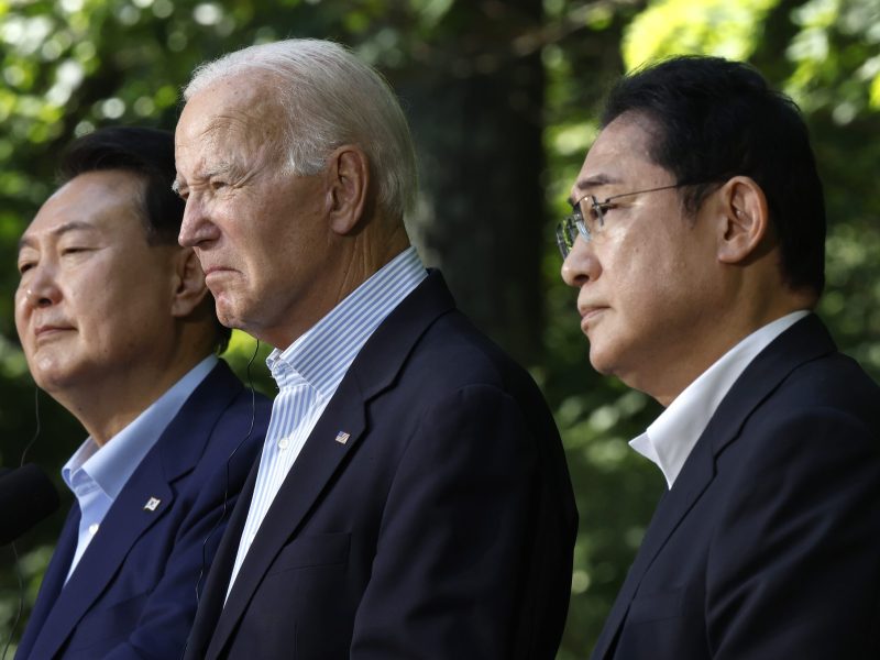 President Yoon, President Biden, and Prime Minister Kishida stand outside together at a press conference in Camp David. They're wearing matching dark suits, light blue shirts, and no ties.