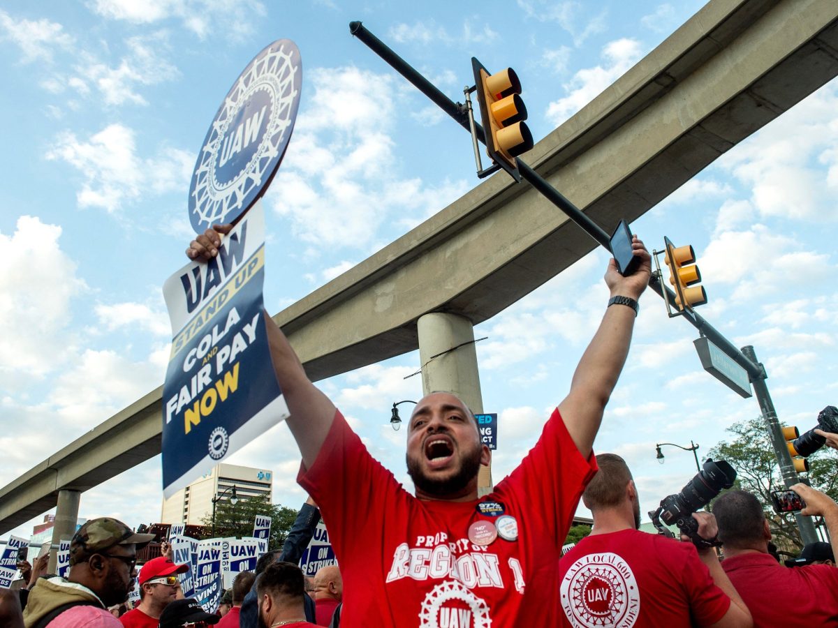 A UAW worker in a red shirt holds up a picket sign with both arms outstretched and a joyous expression on his face. Around him are other red-shirted UAW workers.