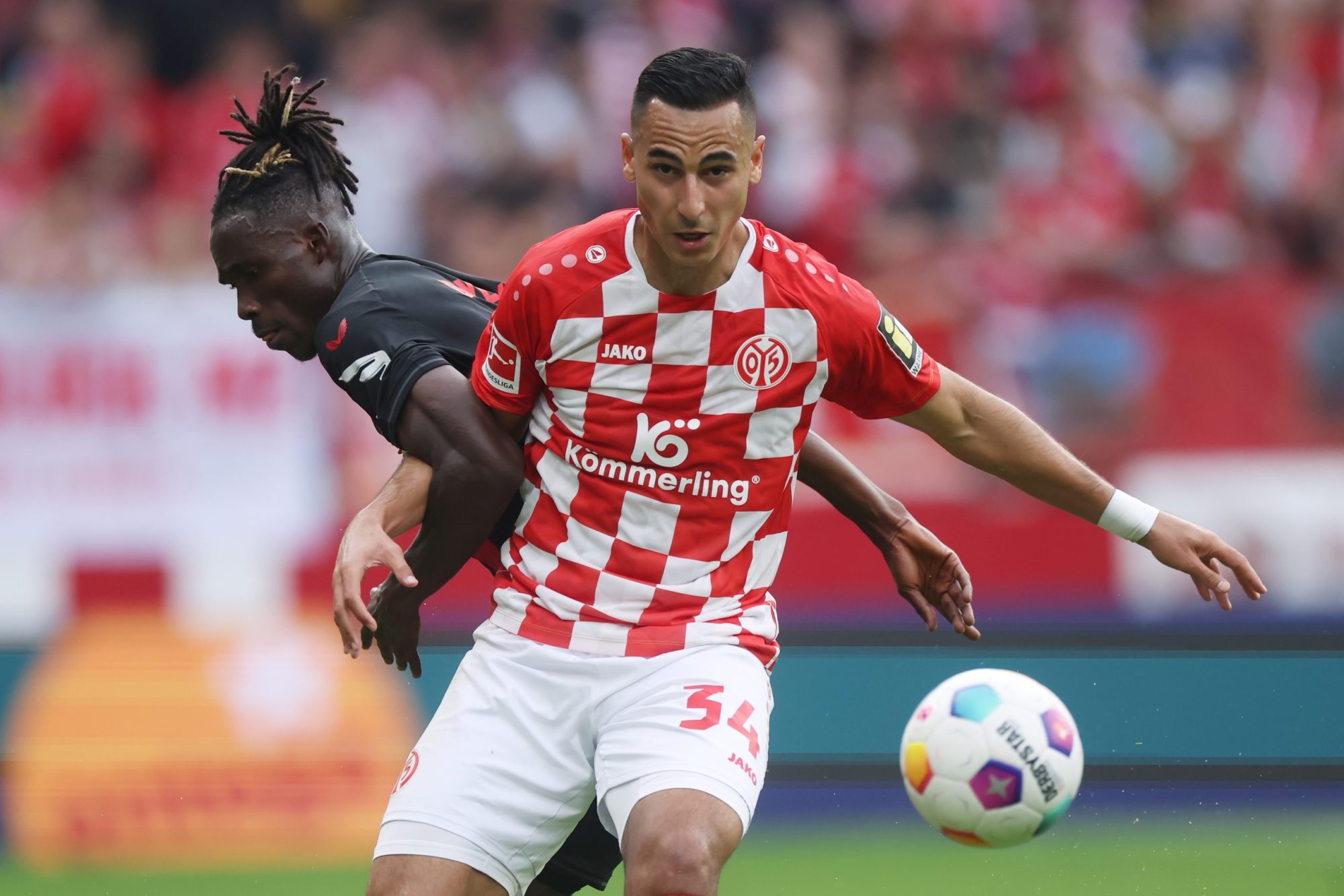 Anwar El Ghazi in the red checkered uniform of the Mainz team, boxing out a challenge on the field from Odilon Kossouneu as the soccer ball flies through the air.