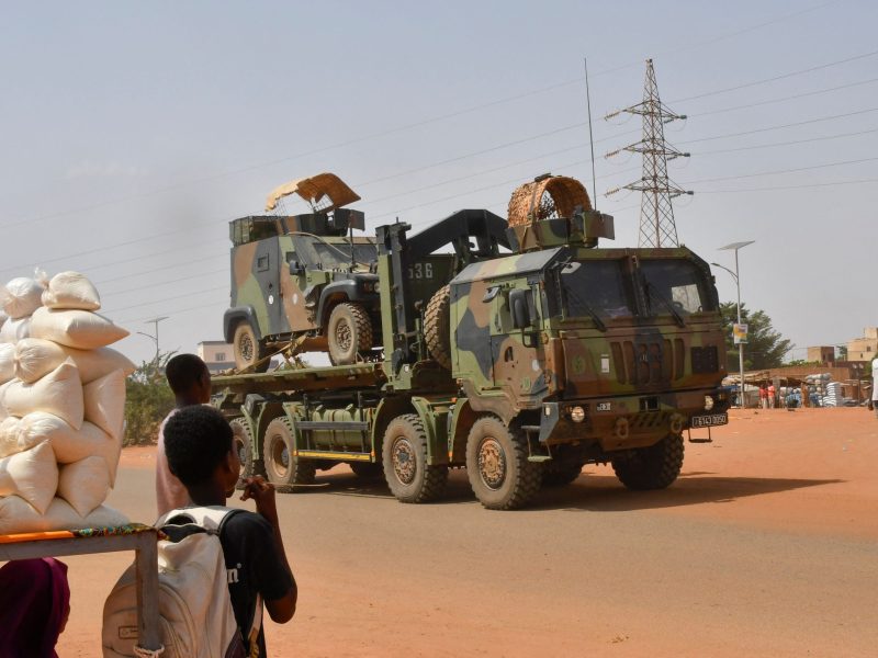 A military supply truck drives off on an unpaved road as two young boys watch.