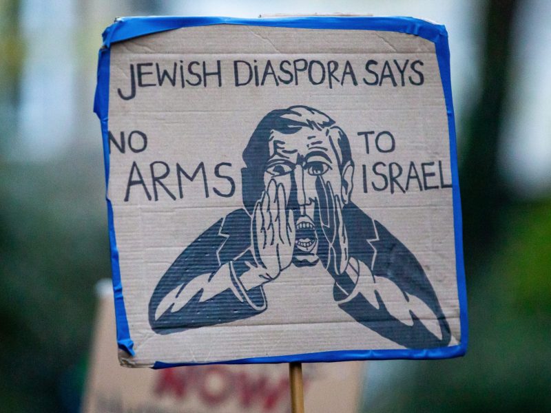 A sign reading 'Jewish Diaspora Says No Arms To Israel'. The sign features an image taken from the Bund, featuring a man with his hands cupping his mouth to amplify something he is saying.