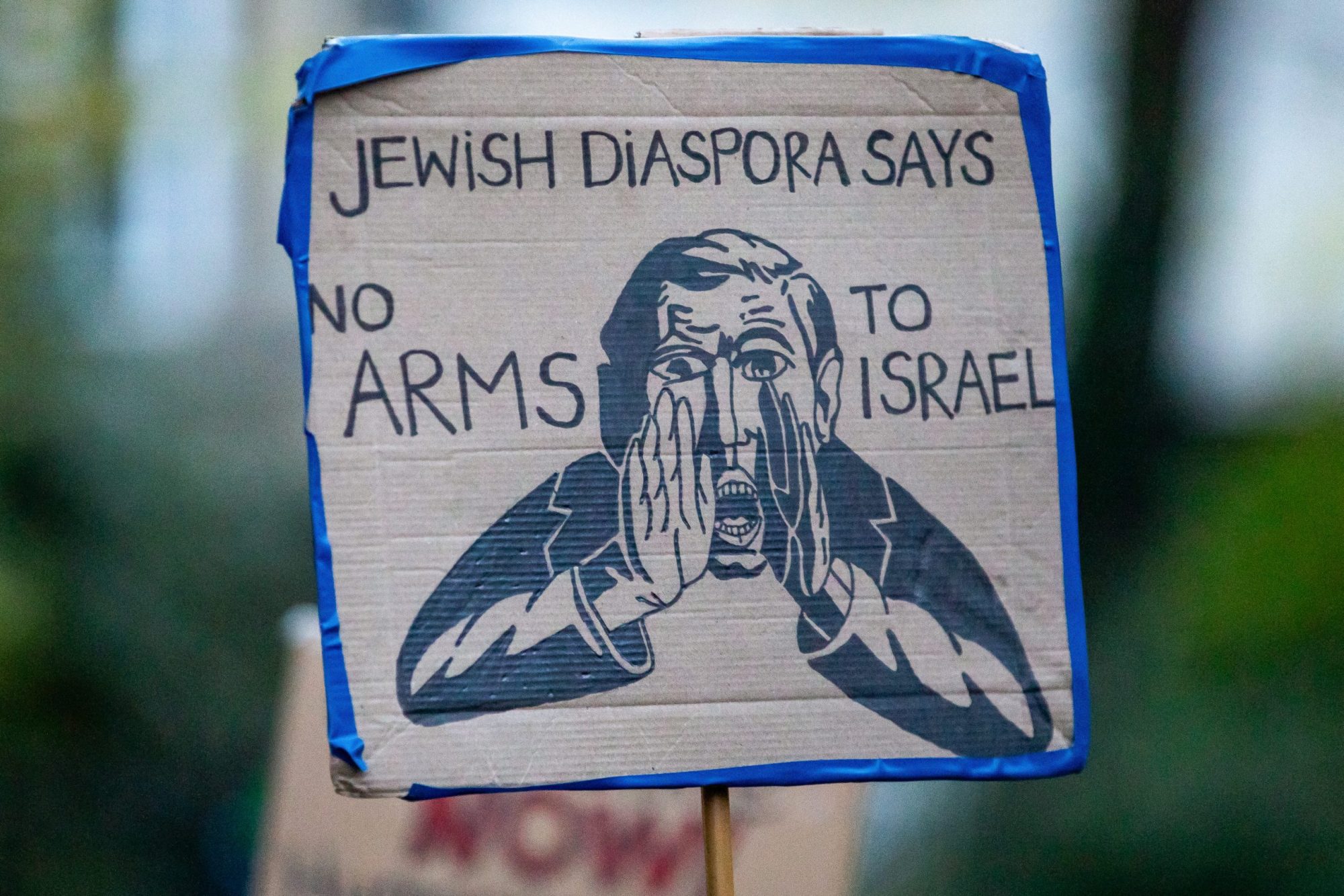 A sign reading 'Jewish Diaspora Says No Arms To Israel'. The sign features an image taken from the Bund, featuring a man with his hands cupping his mouth to amplify something he is saying.