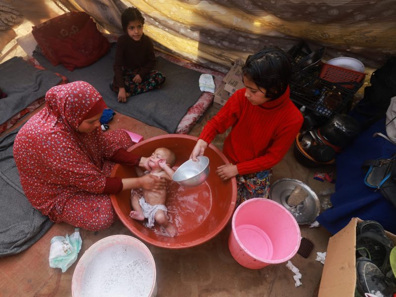 Camera points down at the floor of a tent where thin mats have been spread on top of a tarp. A woman in a red patterned hijab bathes an infant in a red plastic tub on the floor. Her two other daughters sit around her and watch.