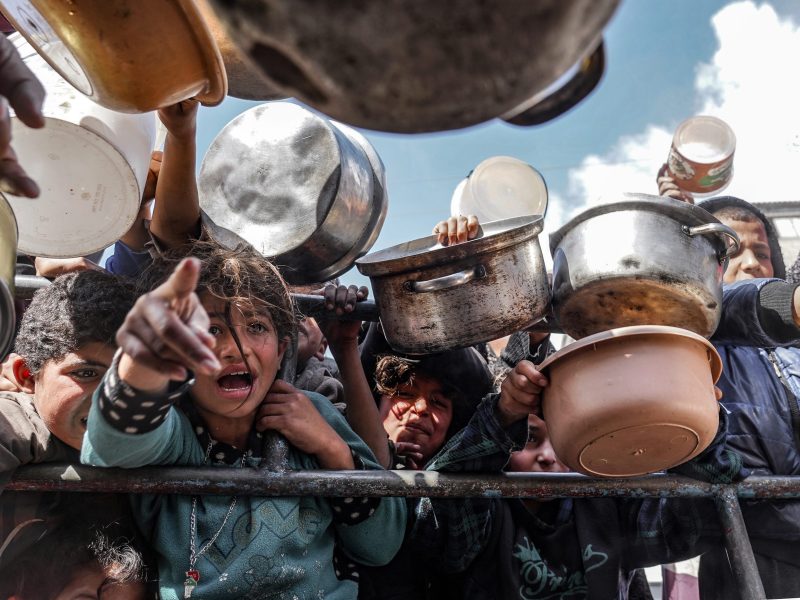 Palestinian children, holding empty pots, wait in line to receive food prepared by volunteers for Palestinian families, displaced to Southern Gaza due to Israeli attacks, between rubble of destroyed buildings in Rafah, Gaza, on Feb. 10, 2024.