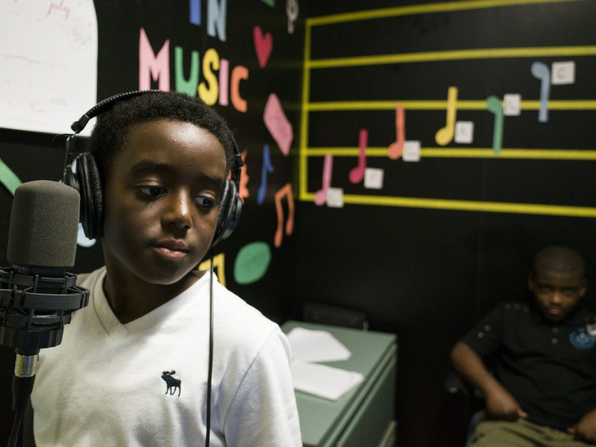 A young Black child wearing a white school uniform and a set of headphones stands in front of a microphone.