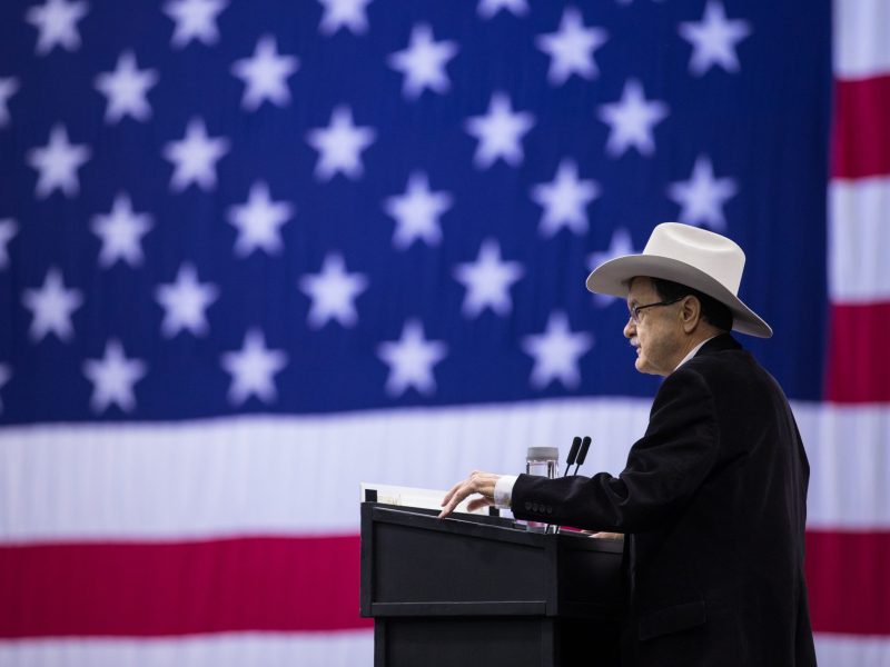 Jim Hightower speaks into a podium. He is wearing a black suit and a cream colored cowboy hat. Behind him is a gigantic American flag.