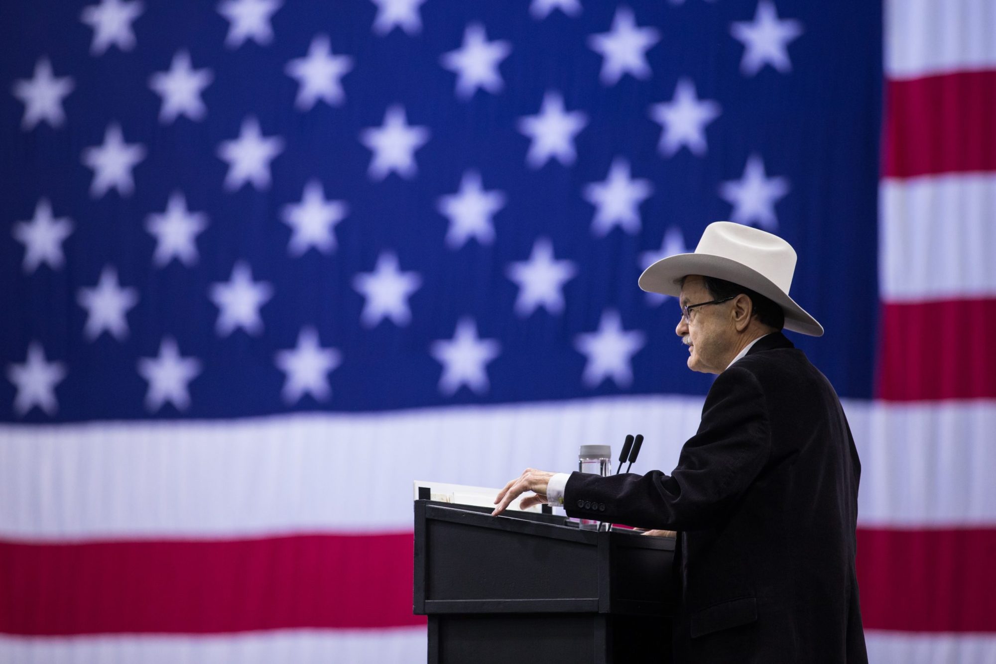Jim Hightower speaks into a podium. He is wearing a black suit and a cream colored cowboy hat. Behind him is a gigantic American flag.