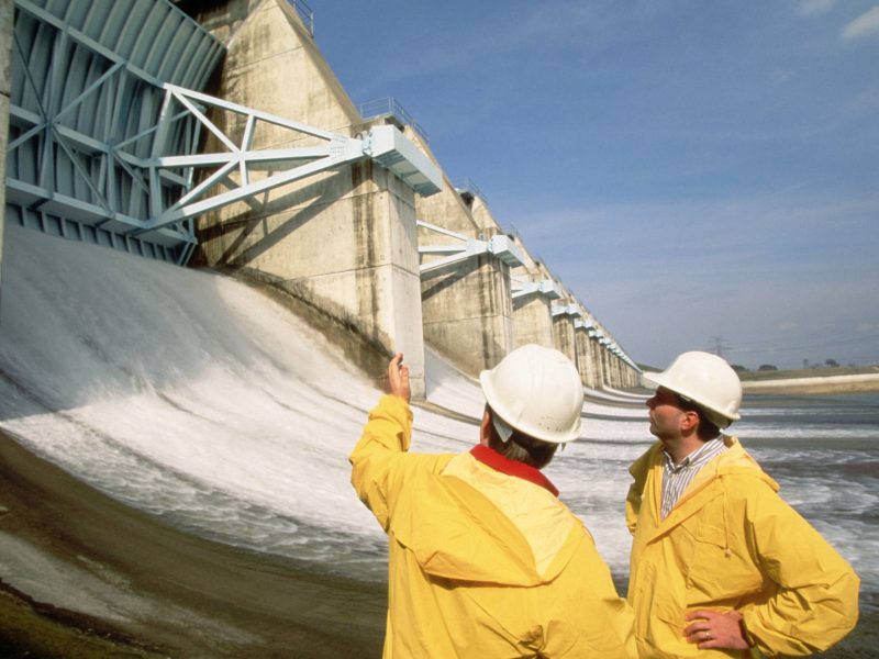 Two people in hardhats and matching yellow utility uniforms stand in front of a dam and point at it, talking.