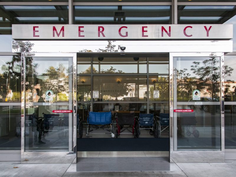 Open sliding glass doors at the entrance to a hospital. The word "Emergency" is written on a sign over the door.