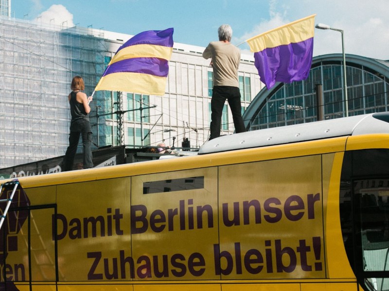 Two activists from Deutsche Wohnen Enteignen wave flags on top of a bus emblazoned with the slogan 'Damit Berlin unser Zuhause bleibt! which translates to 'So that Berlin remains our home!'