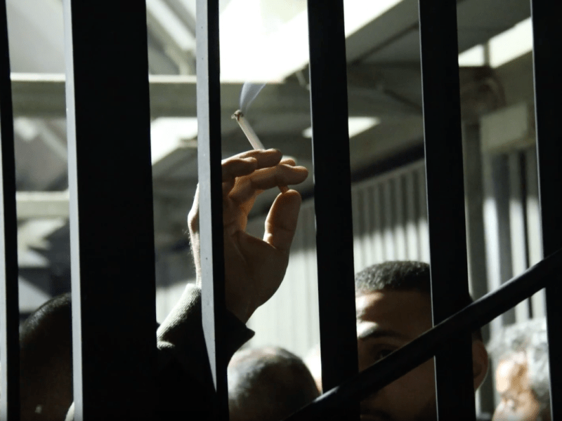 The bars of an Israeli West Bank checkpoint are seen up close. Behind them a Palestinian man's hand holding a cigarette can be seen. The top of his face is visible at the bottom of the photo. Behind him is a harsh fluorescent light, illuminating the darkness.