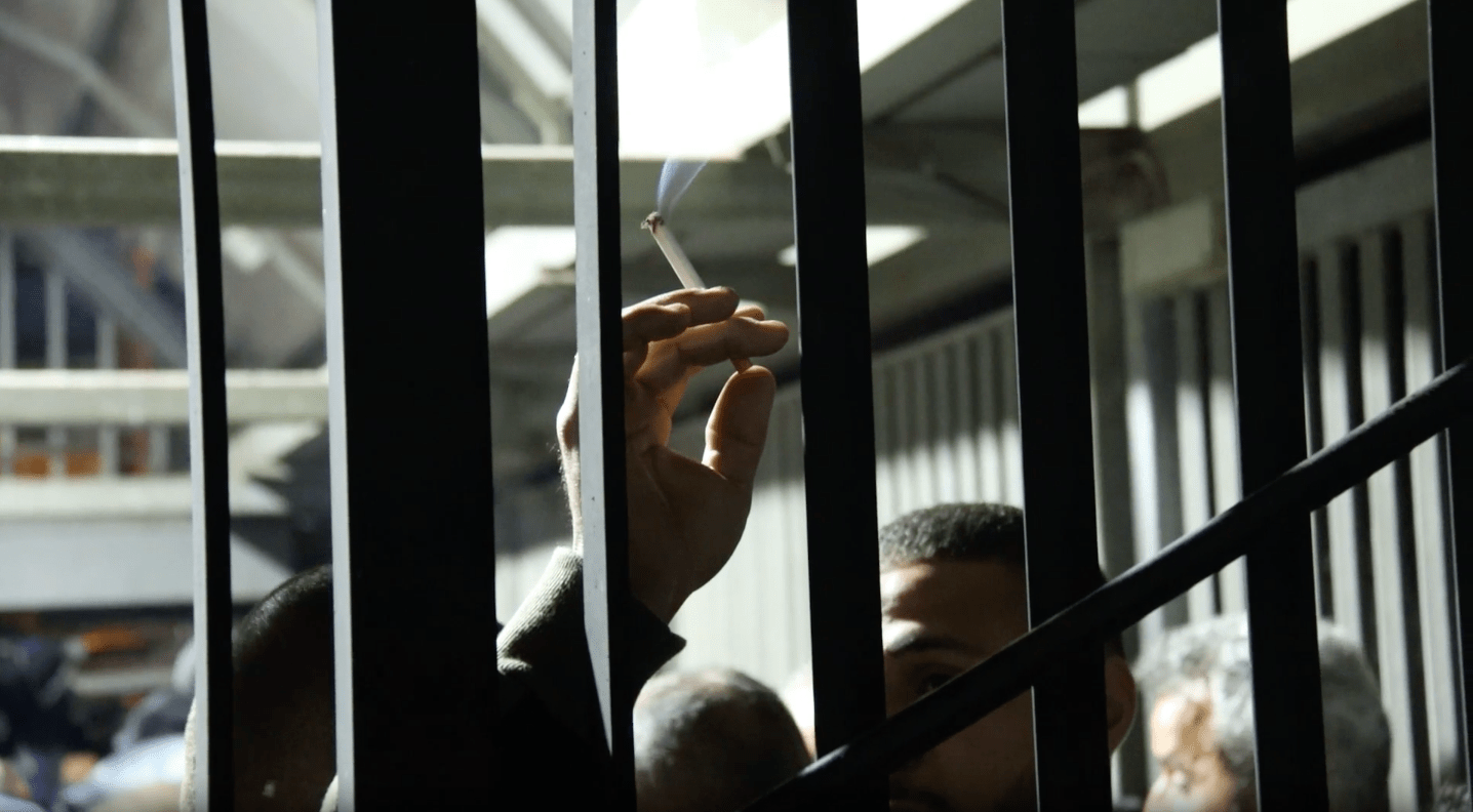 The bars of an Israeli West Bank checkpoint are seen up close. Behind them a Palestinian man's hand holding a cigarette can be seen. The top of his face is visible at the bottom of the photo. Behind him is a harsh fluorescent light, illuminating the darkness.