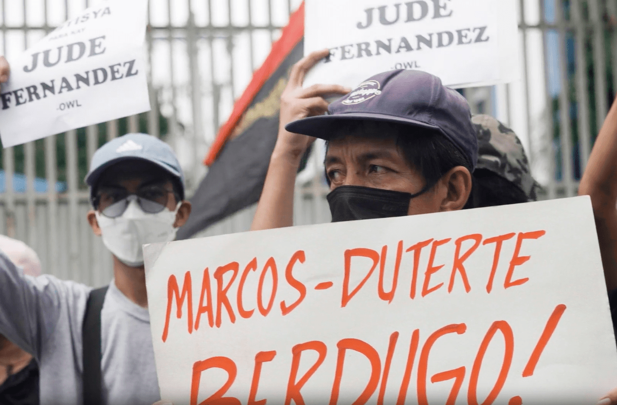 A man wearing a face mask and a hat holds up a sign that says "Marcos-Duterte Berdigo!" In the background two protestors wearing face masks, sunglasses, and caps hold up signs that read "Jude Fernandez"