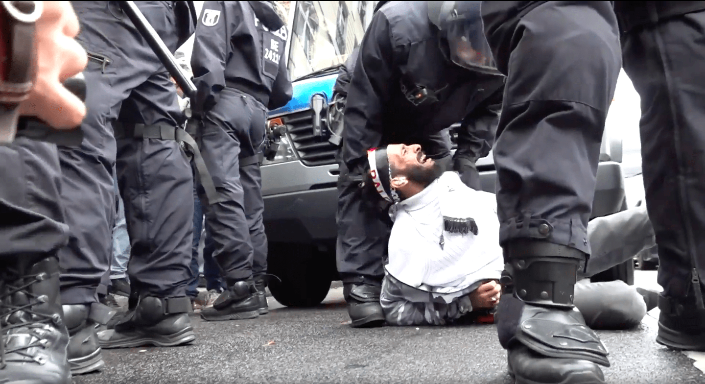 A Middle Eastern man in all white lays down on the pavement with his head rolled back. He is crying out in pain as a black-uniformed police officer bends over to restrain him. In the frame are the black pants and boots of three other cops.